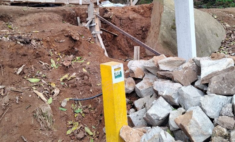 Fotografia que mostra uma área de construção em andamento em meio a uma floresta. Elementos principais da imagem: Uma pilha de rochas de tamanho variado, algumas com marcas de ferramentas, ao lado de um poste amarelo. Uma área de terra escavada ao lado da pilha de rochas, onde se observa a base da construção em andamento. Uma cerca de arame farpado delimitando a área de construção. Árvores e vegetação nativa ao fundo da imagem. Detalhes adicionais: A construção em andamento parece ser de concreto e tijolos. As rochas da pilha podem ter sido utilizadas como base para a construção. A área escavada indica que a construção ainda está em fase inicial. A presença de árvores e vegetação nativa sugere que a área está localizada em um ambiente natural.