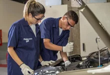 Dois jovens, um rapaz e uma moça, trabalham em um motor de carro. Ele está com uma chave de fenda