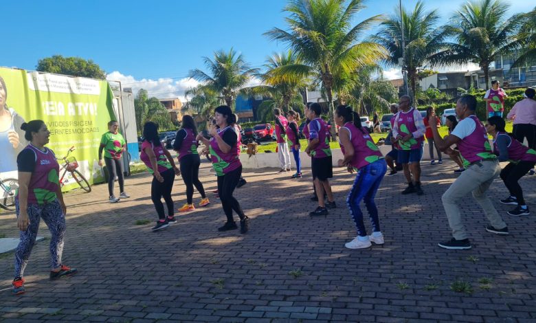 A imagem mostra um grupo de pessoas praticando atividades físicas ao ar livre em uma área com palmeiras e céu claro ao fundo. Os participantes vestem camisetas coloridas e parecem estar envolvidos em uma aula de exercícios, provavelmente uma atividade de ginástica ou dança coletiva. A disposição das pessoas sugere um ambiente animado e colaborativo, com todos realizando movimentos sincronizados. A cena transmite uma atmosfera saudável e ativa, promovendo o bem-estar físico e social em um espaço comunitário.