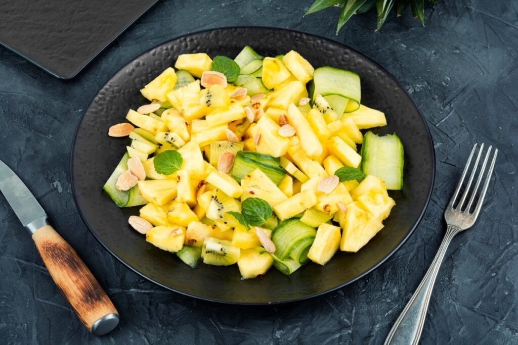 Prato preto com salada de abacaxi, pepino e kiwi decorada com folhas de hortelã 