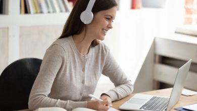 modelo feminino simula, sorridente, estar se preparando para o Enem ouvindo matéria diante de um laptop sobre mesa com livro e caderno de anotação