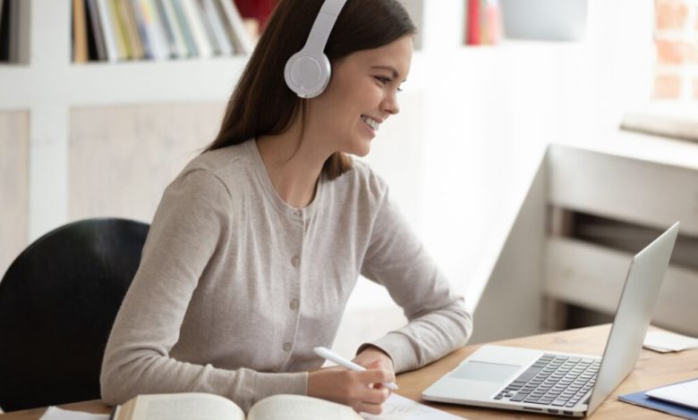 modelo feminino simula, sorridente, estar se preparando para o Enem ouvindo matéria diante de um laptop sobre mesa com livro e caderno de anotação