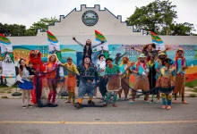 A imagem mostra um grupo de artistas circenses posando em frente a um mural colorido. Eles estão caracterizados com roupas vibrantes e acessórios temáticos, expressando alegria e criatividade. Entre os integrantes, há palhaços, dançarinos e figuras típicas de um circo, segurando redes de pesca e bandeiras que fazem referência ao evento ou projeto "CircoLar". O cenário ao fundo inclui um prédio com o letreiro "AMOCOC" (Associação de Moradores) e murais pintados, retratando uma temática marítima e cultural. A cena transmite um clima festivo e de celebração comunitária.