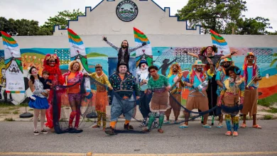 A imagem mostra um grupo de artistas circenses posando em frente a um mural colorido. Eles estão caracterizados com roupas vibrantes e acessórios temáticos, expressando alegria e criatividade. Entre os integrantes, há palhaços, dançarinos e figuras típicas de um circo, segurando redes de pesca e bandeiras que fazem referência ao evento ou projeto "CircoLar". O cenário ao fundo inclui um prédio com o letreiro "AMOCOC" (Associação de Moradores) e murais pintados, retratando uma temática marítima e cultural. A cena transmite um clima festivo e de celebração comunitária.