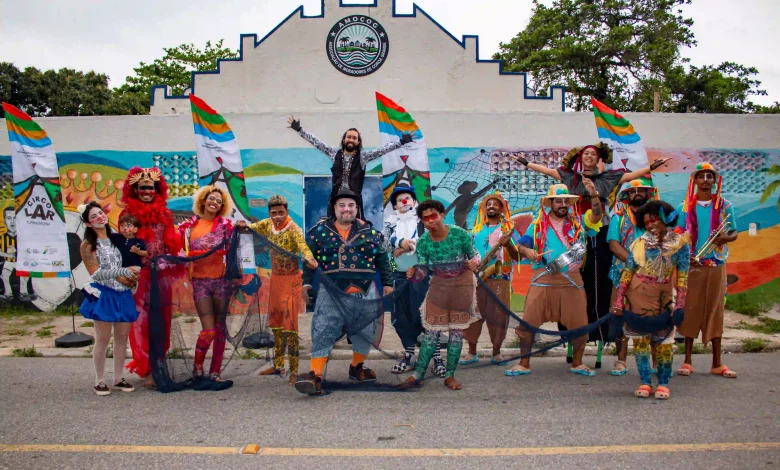 A imagem mostra um grupo de artistas circenses posando em frente a um mural colorido. Eles estão caracterizados com roupas vibrantes e acessórios temáticos, expressando alegria e criatividade. Entre os integrantes, há palhaços, dançarinos e figuras típicas de um circo, segurando redes de pesca e bandeiras que fazem referência ao evento ou projeto "CircoLar". O cenário ao fundo inclui um prédio com o letreiro "AMOCOC" (Associação de Moradores) e murais pintados, retratando uma temática marítima e cultural. A cena transmite um clima festivo e de celebração comunitária.