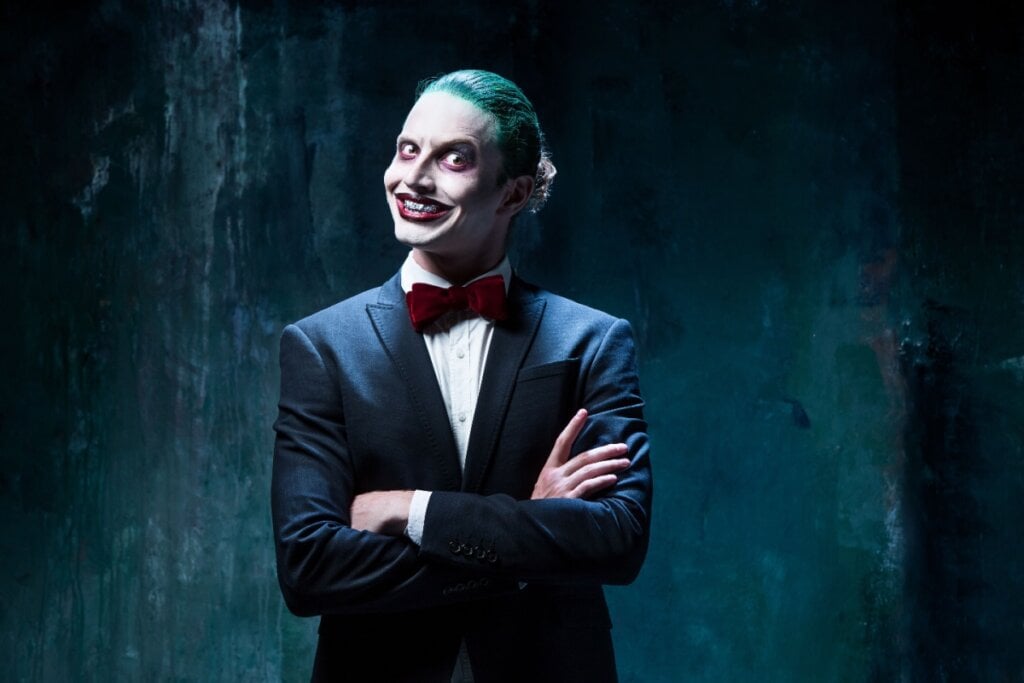 Homem caracterizado como Coringa, com cabelo verde, maquiagem marcante e terno elegante, exibindo um sorriso sinistro em um ambiente escuro