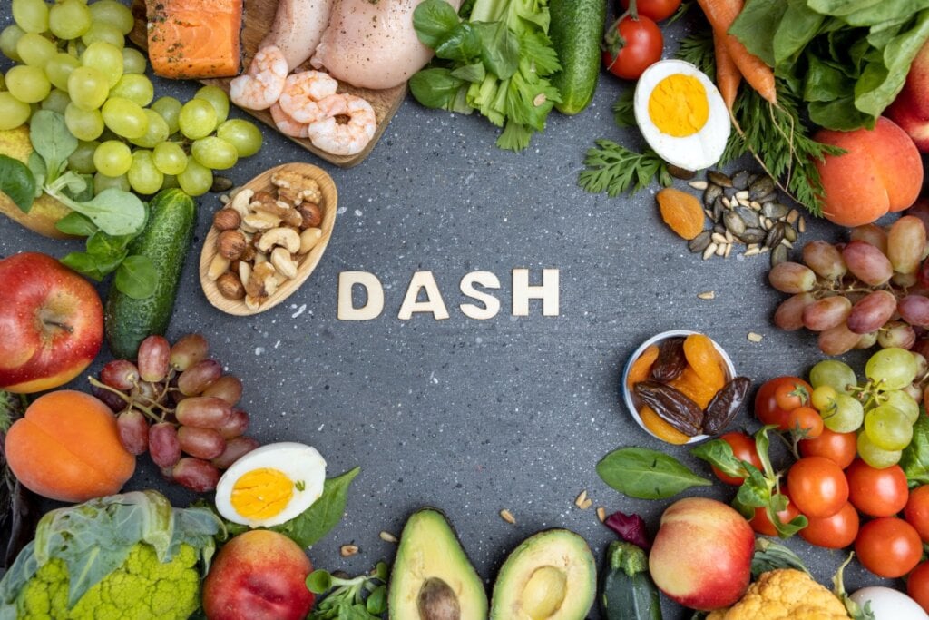 Mesa decorada com alimentos frescos e saudáveis, incluindo frutas, vegetais, nozes, ovos e peixes, dispostos ao redor da palavra "DASH", representando a dieta DASH