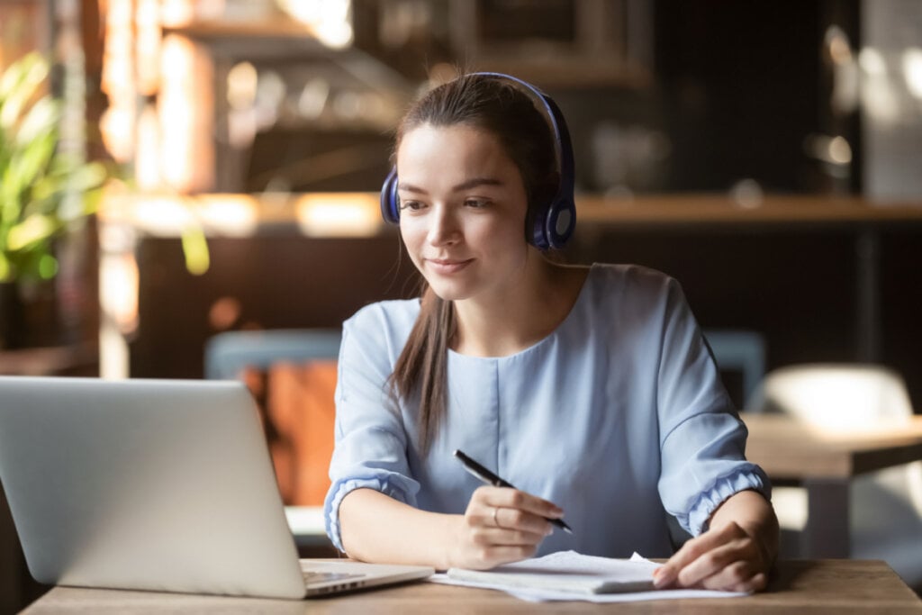 Jovem de fones de ouvido concentrada estudando em ambiente comum com computador e realizando anotações