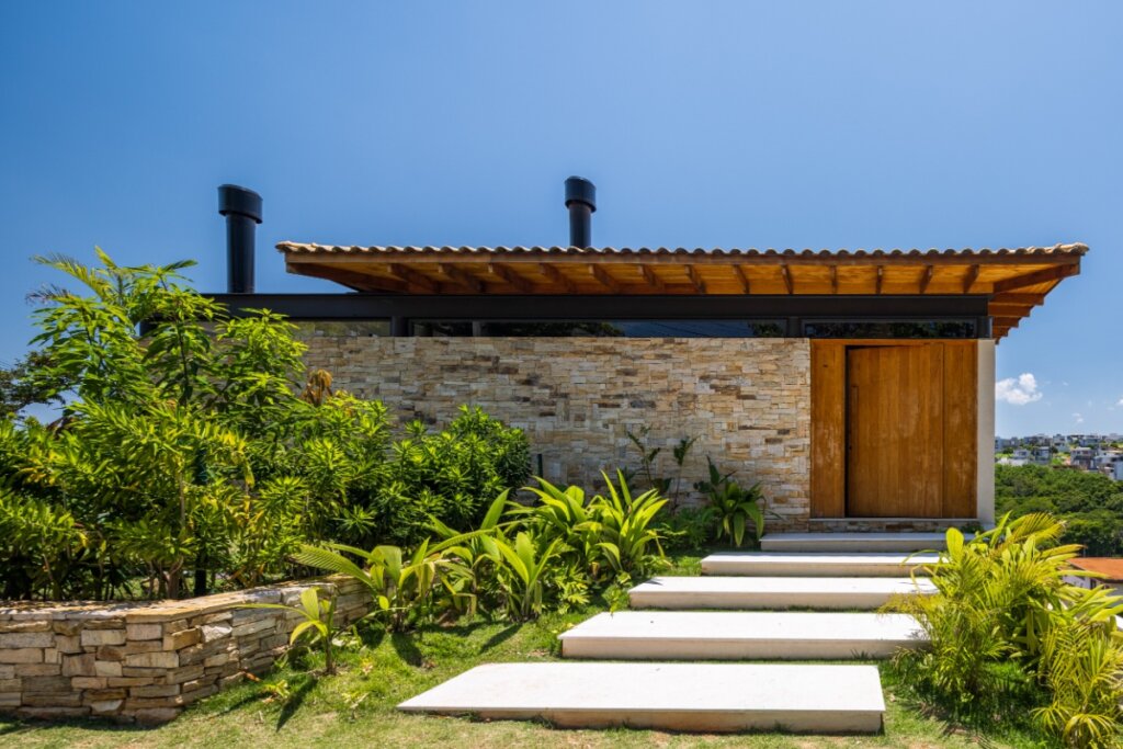 Fachada de casa com porta pivotante de madeira Freijó, integrada ao paisagismo e revestimento em pedras naturais