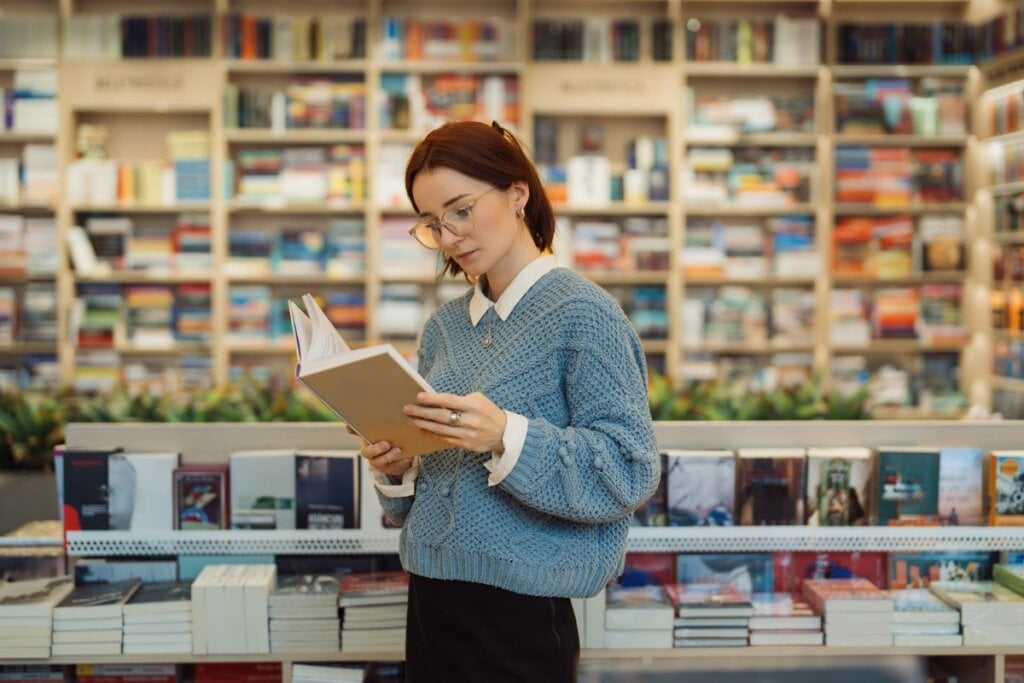 Mulher em pé vestindo uma blusa azul em uma biblioteca lendo livro