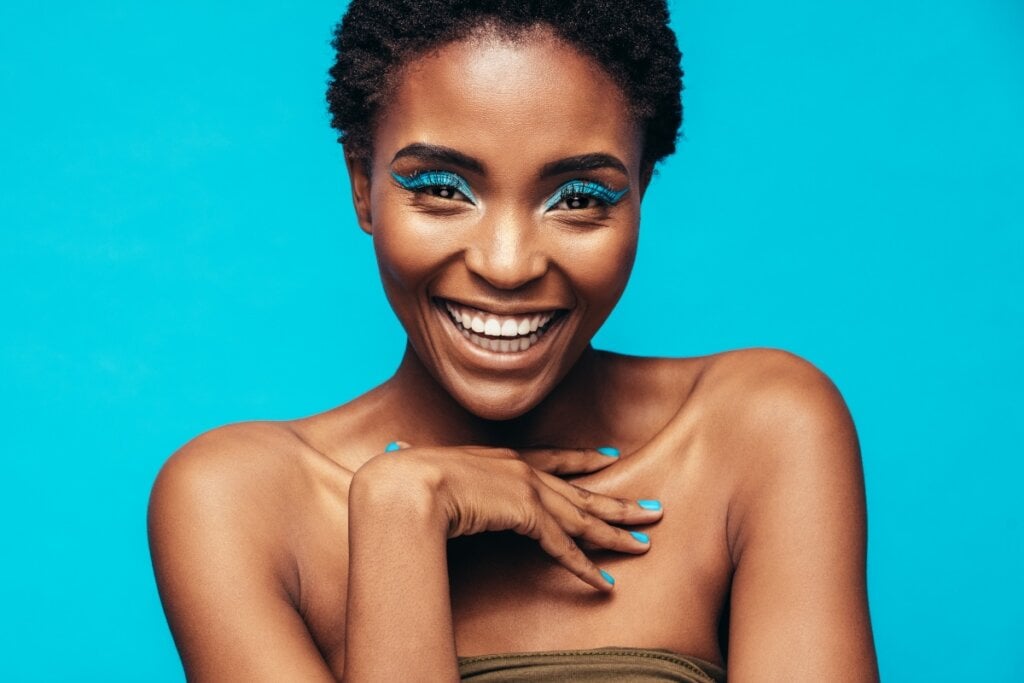 Mulher negra com sombra azul no olho sorrindo com a mão no peito