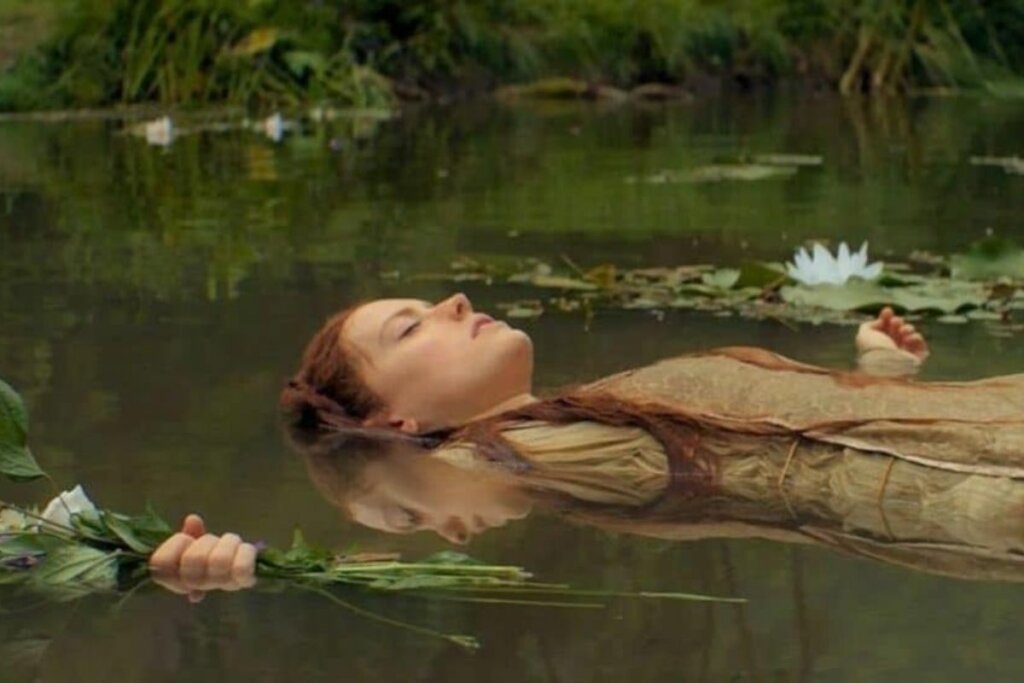 Atriz dentro de um rio com braços abertos e segurando flores em uma cena do filme 'Ophelia'