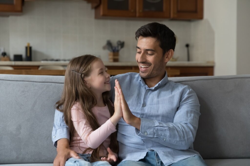 Pai e filha sorrindo em sofá e fazendo "hi-five" com as mãos 