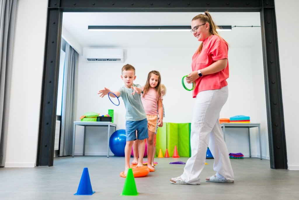 Terapeuta ocupacional guiando duas crianças em um exercício de coordenação motora, utilizando cones coloridos e argolas