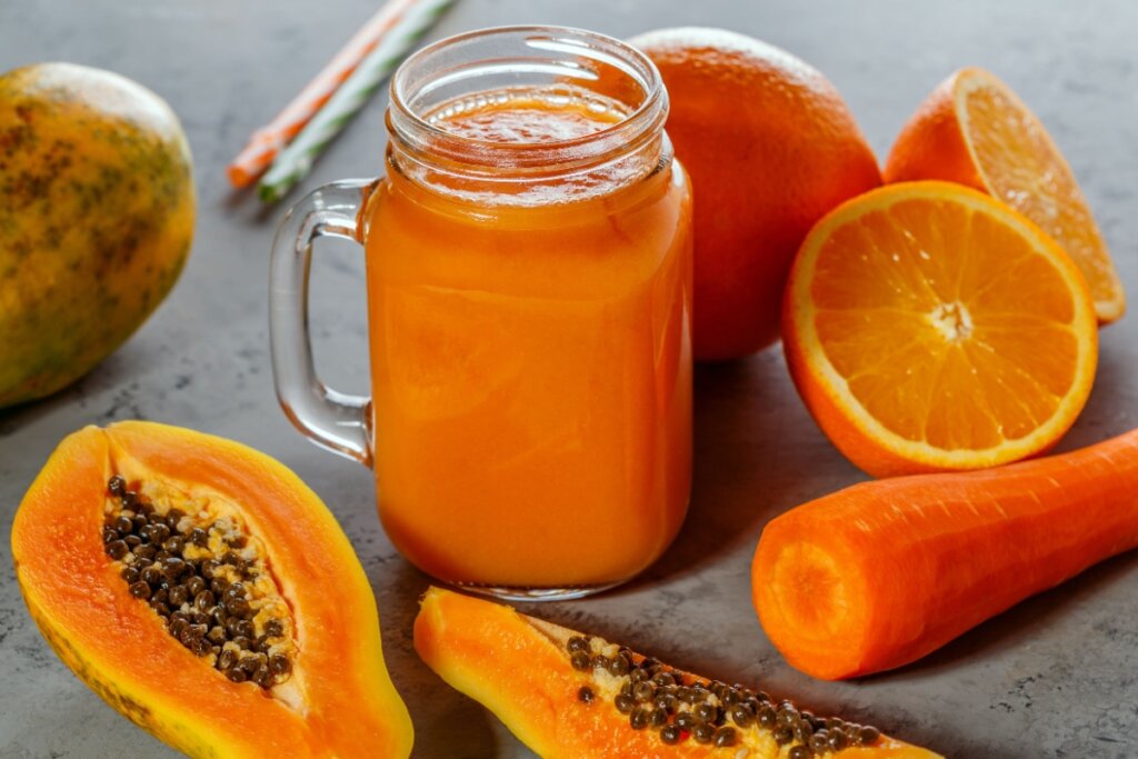 Suco de mamão, laranja e cenoura em copo de vidro; ao lado há mamão, laranja e cenoura cortados