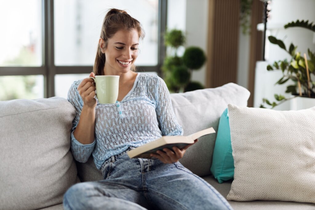 Mulher lendo livro sentada no sofá e bebendo uma caneca de chá