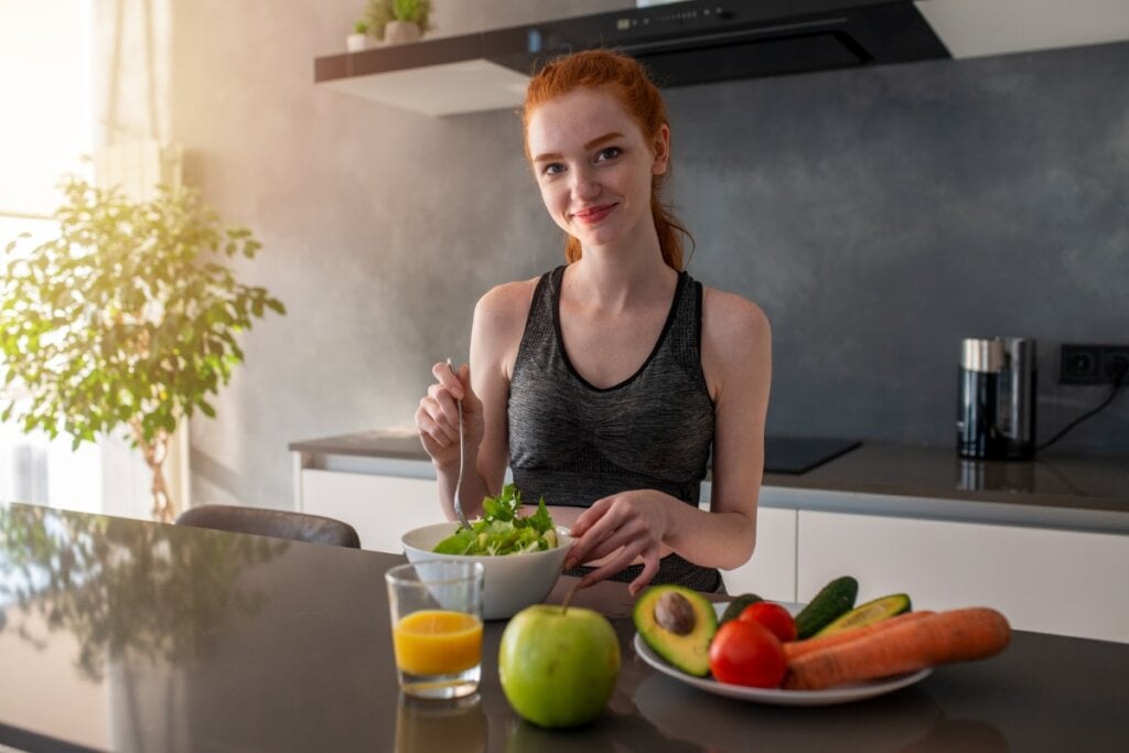 Mulher vestindo um top em uma cozinha em frente a uma mesa com frutas e vegetais