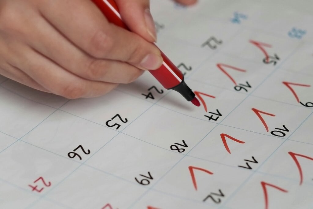 Mão de uma pessoa segurando uma caneta vermelha e fazendo vistos em um calendário 