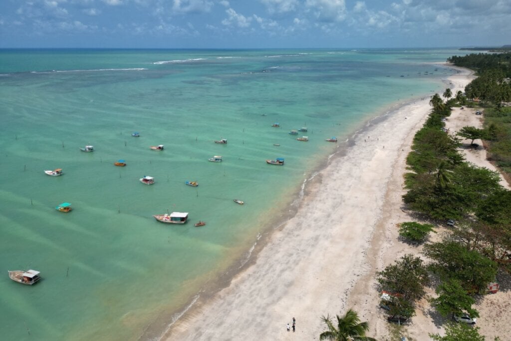 Fotografia aérea da Praia do Patacho, com mar verde-esmeralda, vários barcos ancorados e uma extensa faixa de areia cercada por vegetação