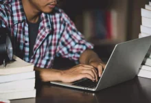 foto mostra jovem mexendo em laptop sbre uma mesa com livros colocados sobre ela