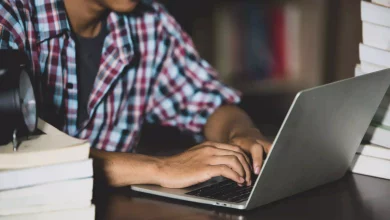 foto mostra jovem mexendo em laptop sbre uma mesa com livros colocados sobre ela