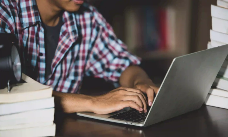 foto mostra jovem mexendo em laptop sbre uma mesa com livros colocados sobre ela