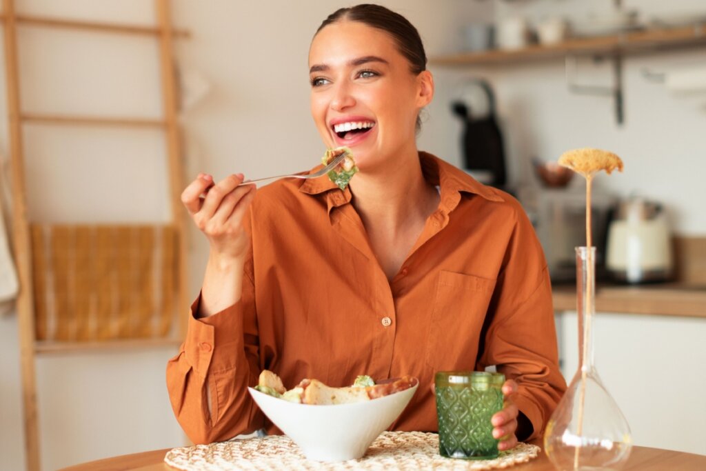 mulher de blusa marrom, sentada e levando garfo com salada à boca