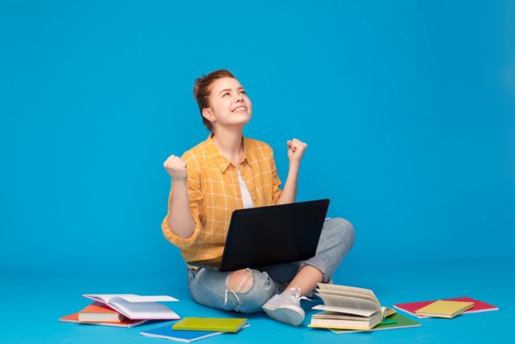 Estudante comemorando com expressão de felicidade, cercada por livros e um notebook