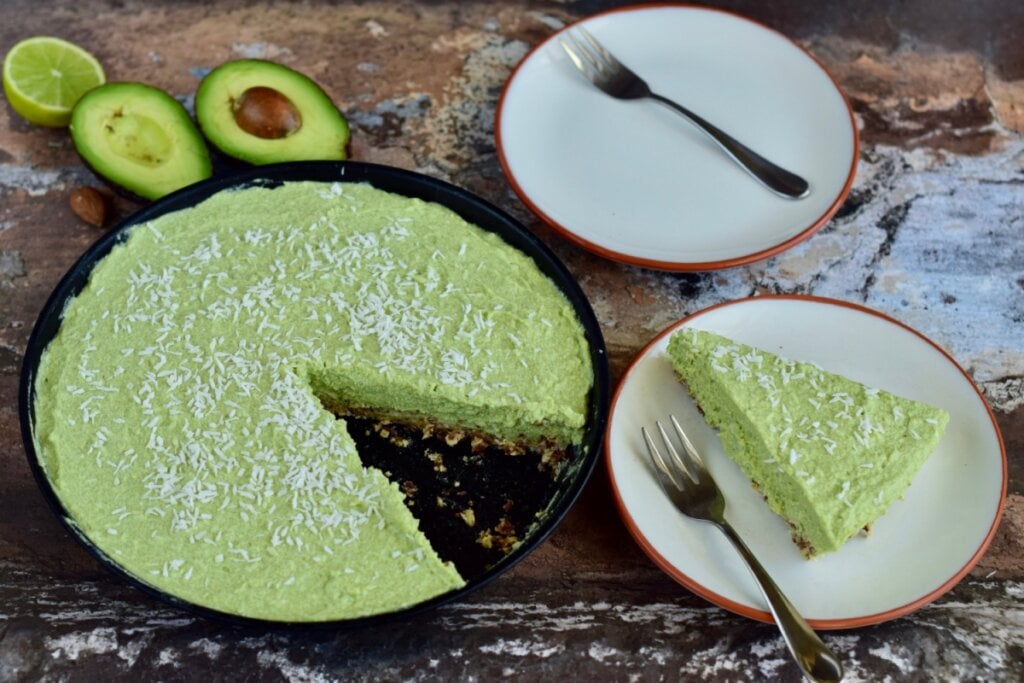 Torta gelada de abacate servida em cima de mesa de madeira. Ao lado um prato branco com um pedaço de torta e um garfo de sobremesa e ao lado também há outro prato branco vazio com um garfo