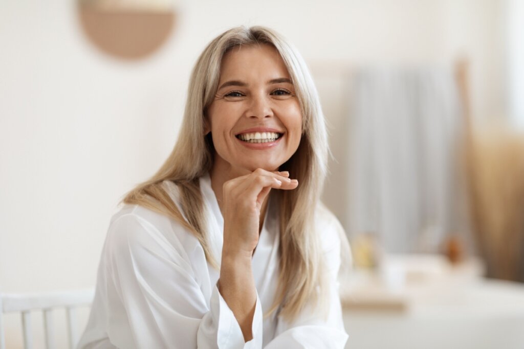 Mulher sorrindo, usando um roupão branco e sentada com a mão apoiada no queixo 