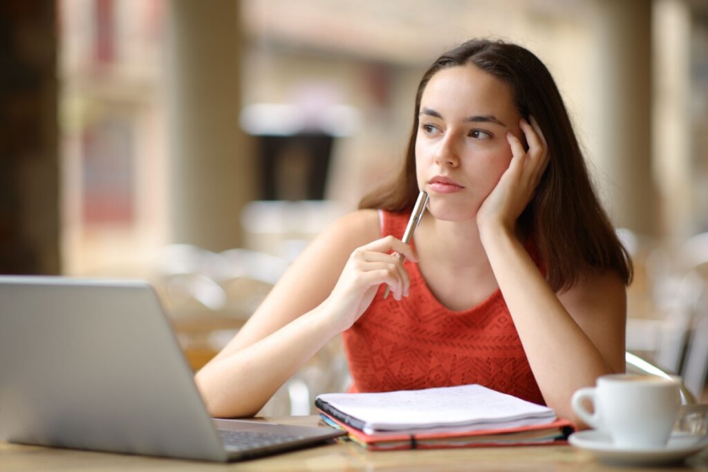 Estudante, sentada em frente a uma mesa com notebook e papéis, pensando 