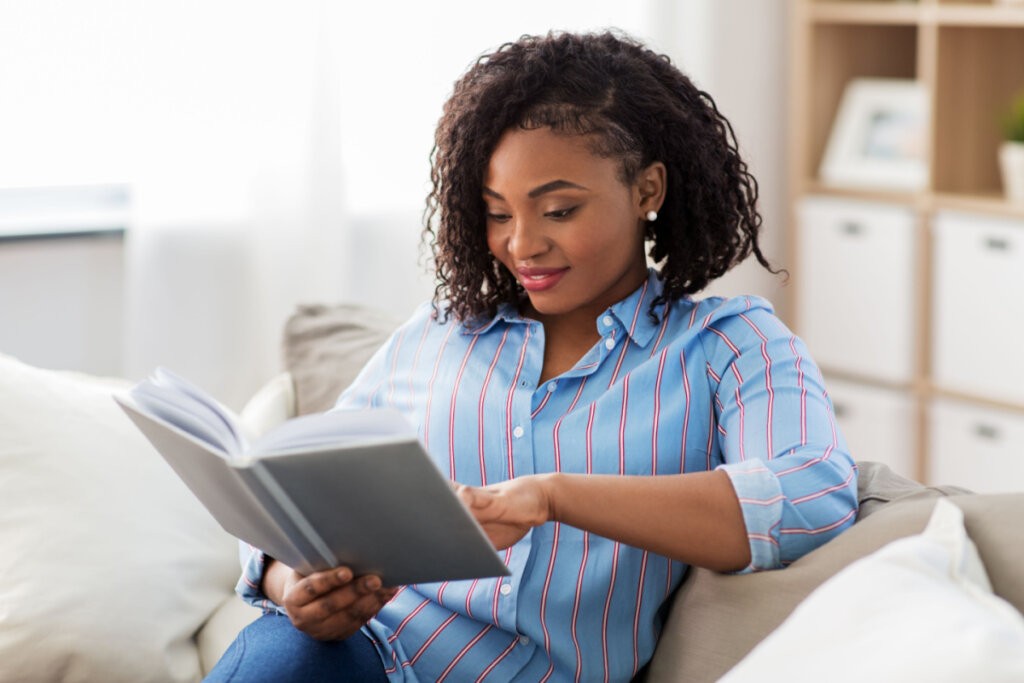 Mulher de blusa azul, sentada no sofá e lendo livro de capa cinza