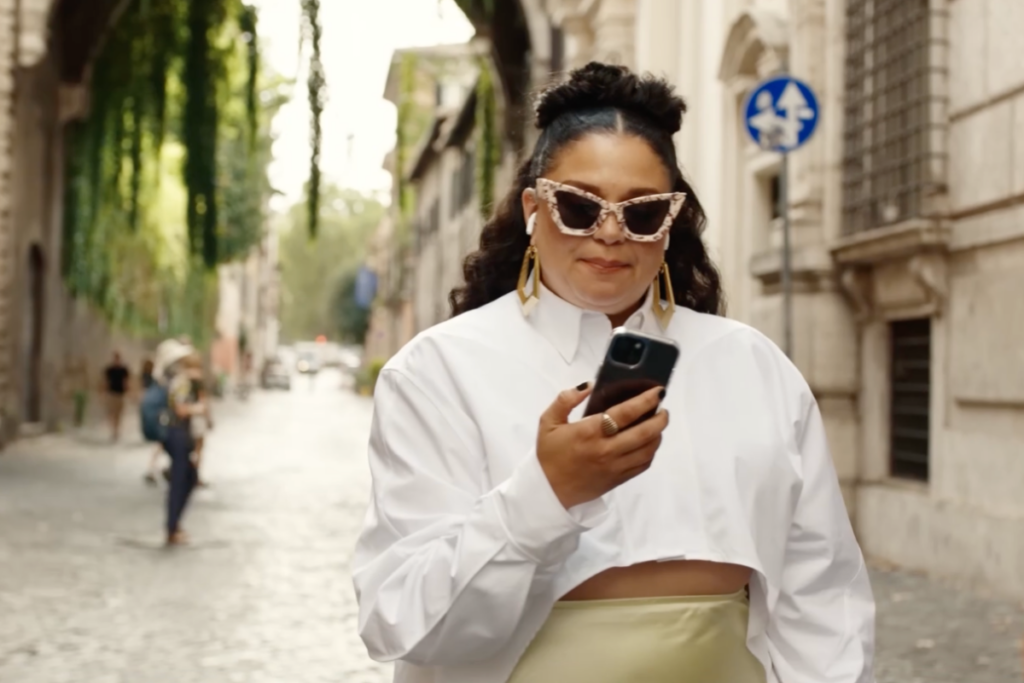 Mulher estilosa caminhando em uma rua movimentada, de óculos escuros e celular na mão