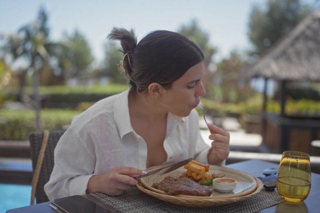 Mulher sentada de frente  para uma vista com piscina comendo um bife 