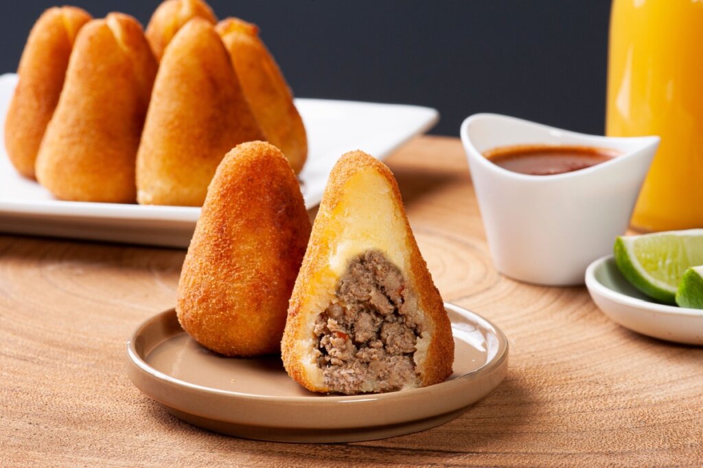 Coxinha de carne moída, com massa dourada e macia, servida em um prato acompanhado de molho, limão e suco