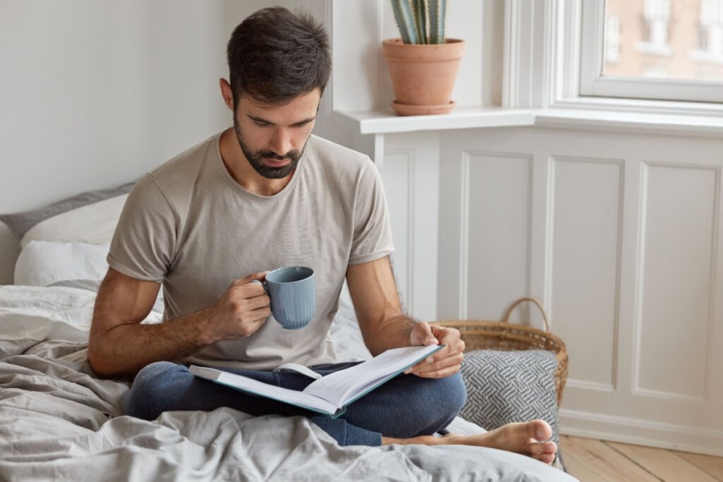 homem lendo livro segurando caneca sentado em uma cama