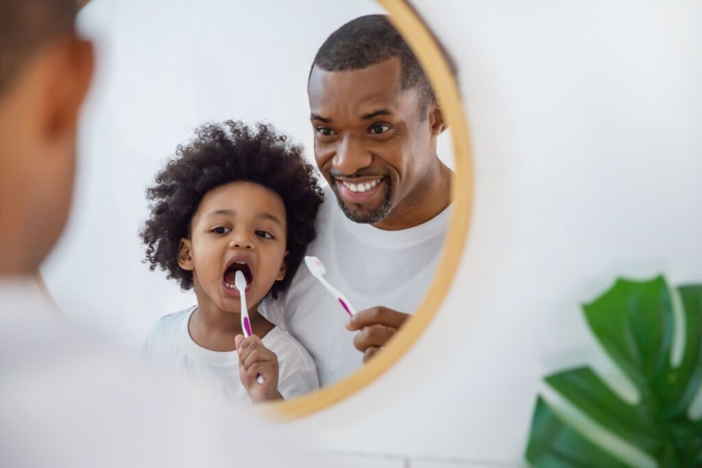 Menino em frente a um espelho escovando os dentes e um homem do lado olhando e sorrindo 