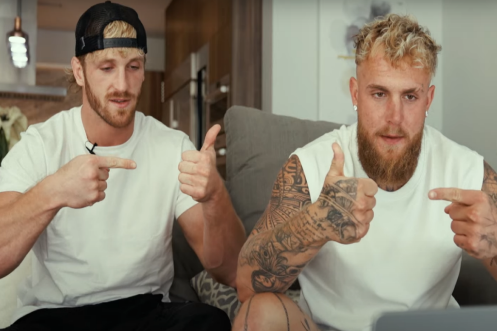 Imagem de dois homens vendo e imitando vídeo, os dois estão usando camisetas brancas e um deles usa um boné preto