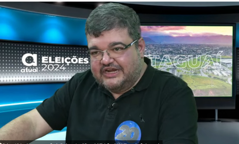 Rubão, de óculos e blusa preta, tendo ao fundo um cenário com a logomarca do jornal atual com a inscrição Eleições 2024 à esquerda e Itaguaí à direita