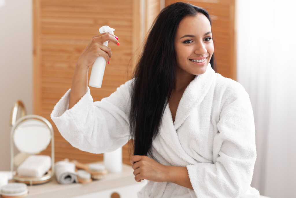 Mulher em um banheiro, usando um roupão branco e passando um produto no cabelo 