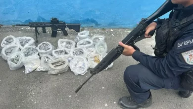 Policial Militar agachado diante de um fuzil e farta quantidade de drogas apreendidos