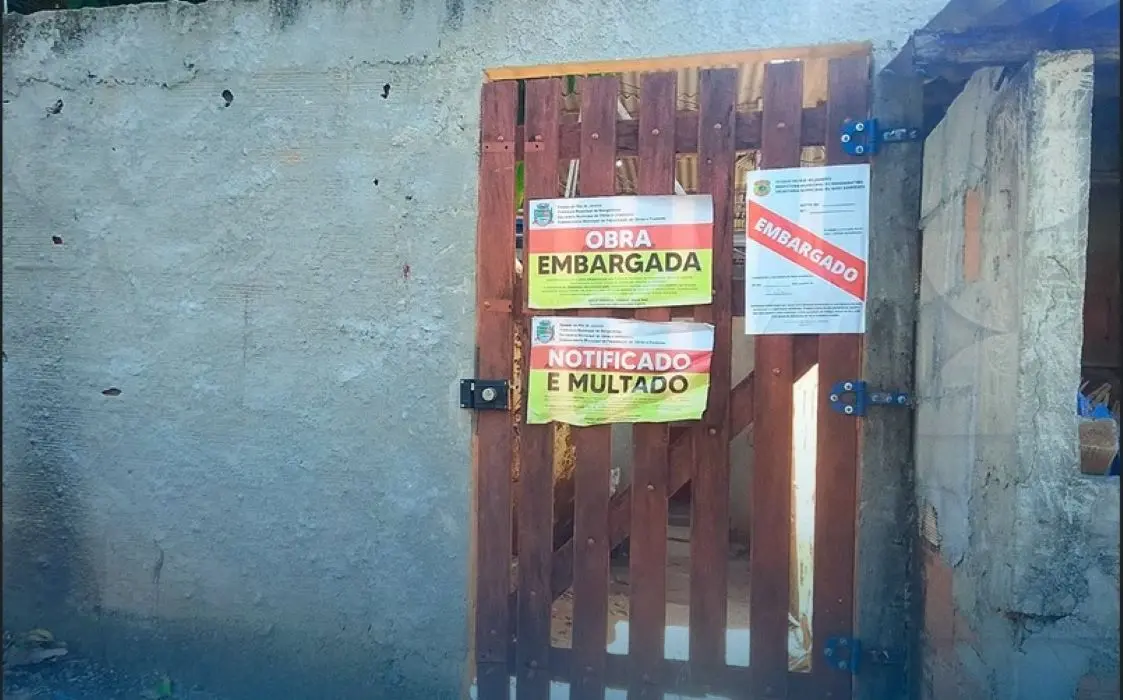 Avisos de "Obra Embargada" e "Notificado e Multado" são afixados em portão de construção irregular