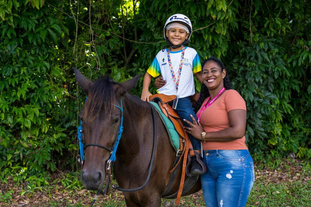 Criança em cima do cavalo com a professora em pé ao lado durante a equoterapia