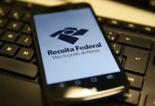 Celular com tela ligada no aplicativo da Receita Federal está deitado em cima de um teclado de computador.