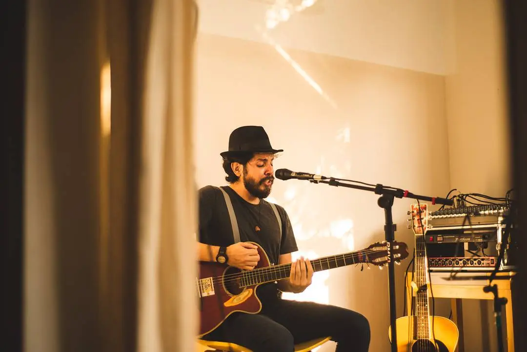 Homem branco usando roupa preta está sentando tocando violão