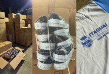Foto mostra caixas de papelão empilhadas em galpão, pares de tênis pretos com solado branco identificados com o logotipo “Itaguaí Prefeitura” e uma camiseta cinza da rede municipal com o brasão do município e a palavra “Educação”.