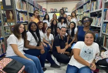 Grupo de jovens posa para a foto junto a professores, numa sala com estantes cheias de livros