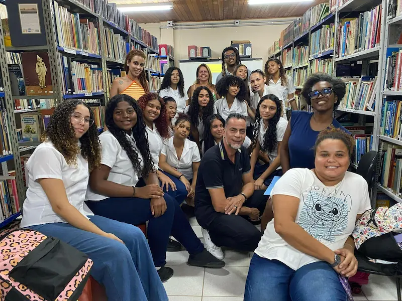 Grupo de jovens posa para a foto junto a professores, numa sala com estantes cheias de livros