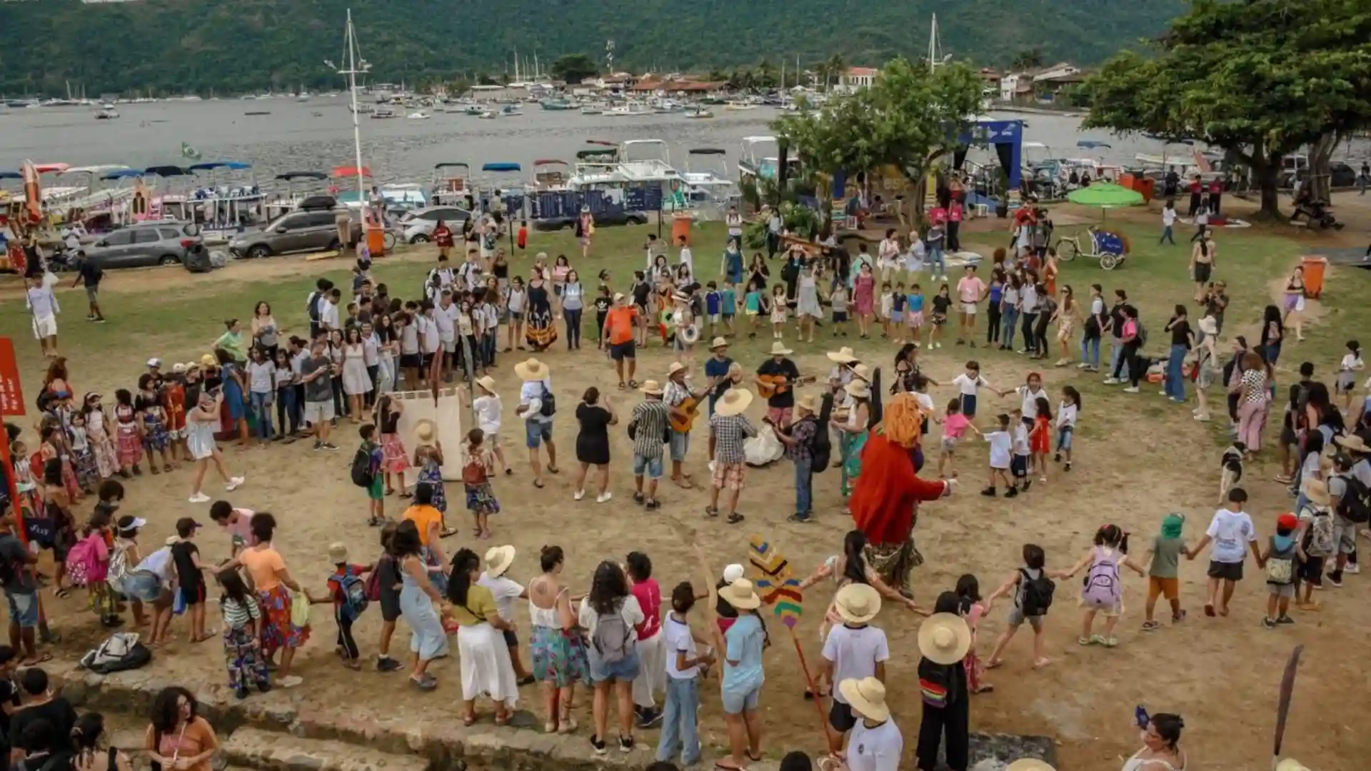 Roda formada por dezenas de pessoas — crianças, jovens e adultos — participando de atividades culturais ao ar livre em Paraty, com o mar e barcos ao fundo, durante o 1º Festival de Cultura Caiçara da Costa Verde.