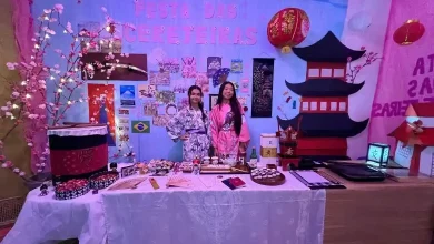 Duas estudantes vestidas com quimonos posam em frente a um estande temático da cultura japonesa, com decoração de cerejeiras, lanternas, maquete de pagode e mesa com objetos e comidas típicas, durante a Feira Mundo do Colégio Educandário São José REALIZADA EM 2024